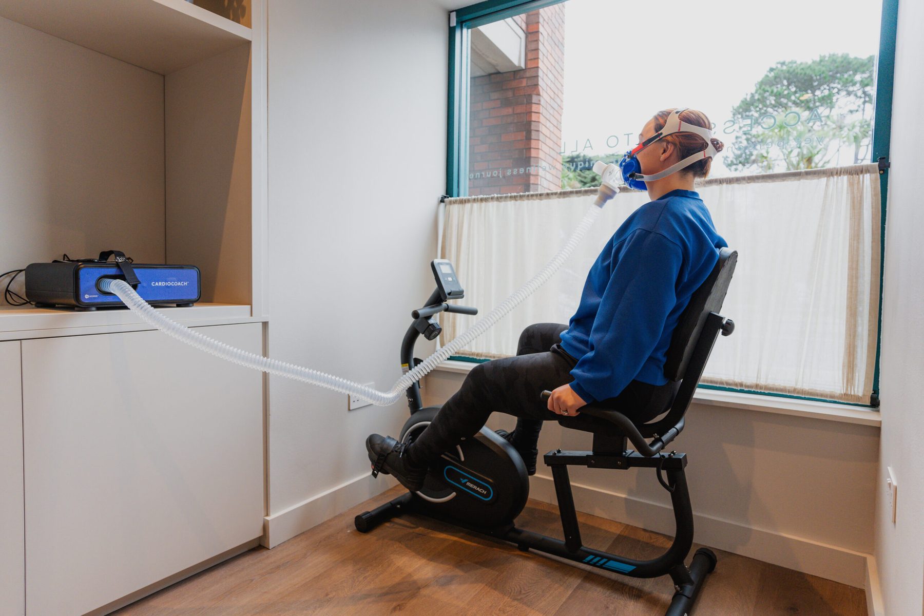 A man seated on exercise equipment by a window wearing a gas exchange mask during a test